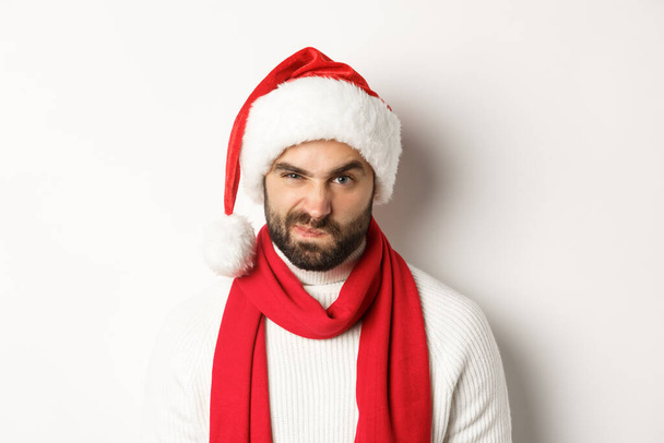 Nieuwjaarsfeest en wintervakantie concept. close-up van chagrijnige kerel in santa hoed fronsen en grimmig, staande tegen witte achtergrond - Foto, afbeelding