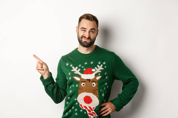 Újévi ünneplés és téli ünnepek koncepció. Szkeptikus srác karácsonyi pulóver néz elégedetlen, ujjal mutogatott a bal felső sarokban termék, nem tetszik, fehér háttér - Fotó, kép