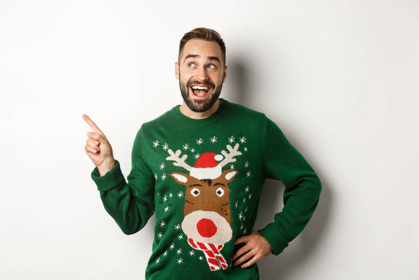 Winterurlaub und Weihnachten. Glücklicher Mann im lustigen Pullover, der mit dem Finger auf das Logo oben links zeigt, zeigt Werbung, weißer Hintergrund - Foto, Bild