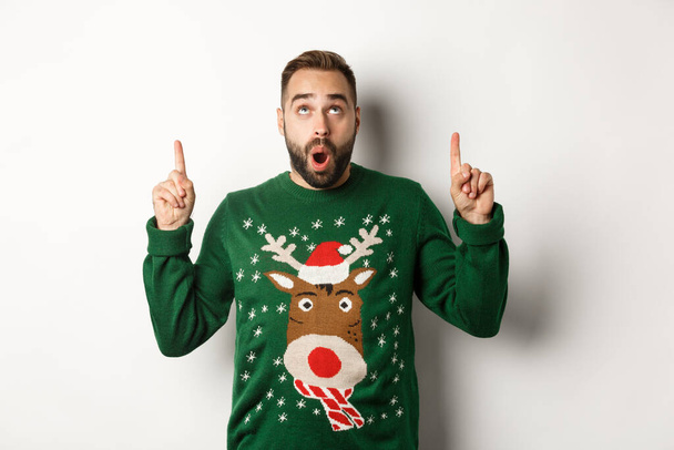 Téli ünnepek és karácsony. Lenyűgöző jóképű férfi újévi pulóverben ujjal felfelé, logót mutat, fehér háttér felett áll - Fotó, kép