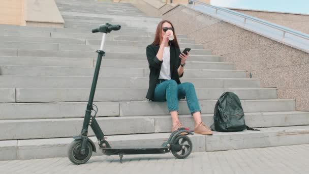 Teljes lövés a fiatal vonzó vegyes verseny nő ül a beton lépcsőn a szabadban kortyolgatva kávét és szörfözés háló az ő okostelefon mosolygós elektromos robogó előtérben - Felvétel, videó