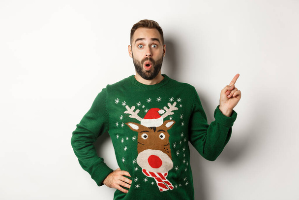 Winterurlaub und Weihnachten. Mann schaut aufgeregt, als er mit dem Finger direkt auf Logo zeigt, steht erstaunt vor weißem Hintergrund - Foto, Bild