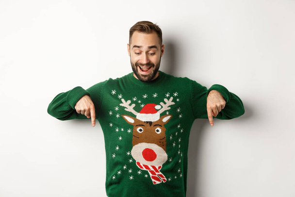 Zimní prázdniny a Vánoce. Šťastný vousatý muž v zeleném svetru, dívá se a ukazuje prsty dolů na reklamu, ukazuje kopírovací prostor, bílé pozadí - Fotografie, Obrázek