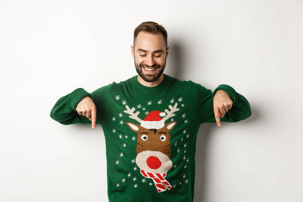 Téli ünnepek és karácsony. Boldog fehér férfi zöld pulóverben, ujjal lefelé mutogatva és elégedetten, fehér háttér felett állva - Fotó, kép