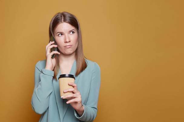 Serious woman entrepreneur poses with takeaway coffee, smartphone, works on new business project, has important conversation, dressed in formal outfit, poses agains yellow background, checks schedule. - Фото, изображение