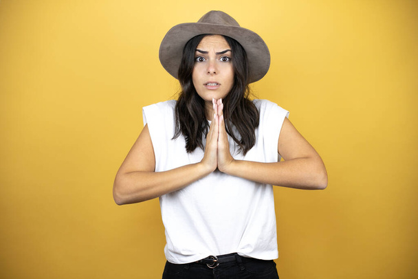 Beautiful woman wearing casual white t-shirt and a hat standing over yellow background begging and praying with hands together with hope expression on face very emotional and worried - Photo, Image