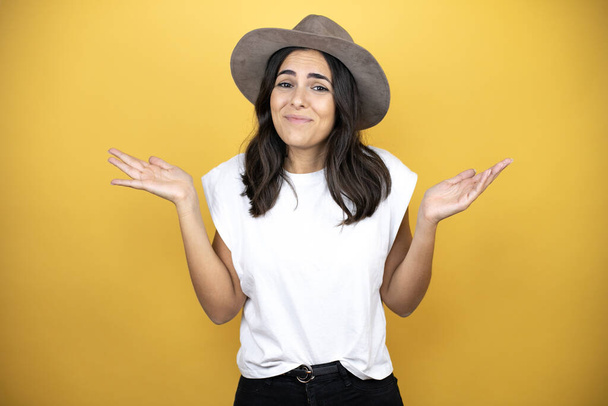 Beautiful woman wearing casual white t-shirt and a hat standing over yellow background clueless and confused expression with arms and hands raised - Photo, Image