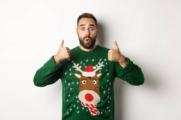 Weihnachten, Feiertage und Feiern. Überraschter junger Mann in grünem Pullover, Daumen hoch in Anerkennung, wie etwas Großes, vor weißem Hintergrund stehend - Foto, Bild