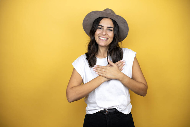 Schöne Frau mit legerem weißen T-Shirt und Hut vor gelbem Hintergrund, lächelnd mit den Händen auf der Brust und dankbarer Geste im Gesicht. - Foto, Bild