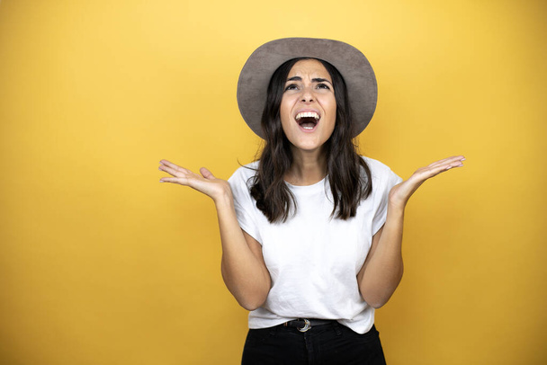 Beautiful woman wearing casual white t-shirt and a hat standing over yellow background clueless and confused expression with arms and hands raised - Valokuva, kuva