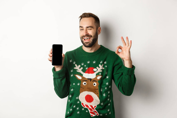 Christmas holidays and shopping concept. Confident guy winking and showing okay sign with mobile screen, standing over white background - Photo, Image