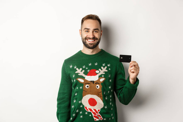 Christmas holidays and shopping concept. Young man in winter sweater showing credit card, standing over white background - Fotó, kép