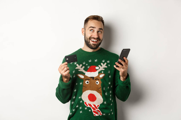 New Year, online shopping and christmas concept. Young man buying gifts in internet, holding mobile phone and credit card, white background - Fotografie, Obrázek