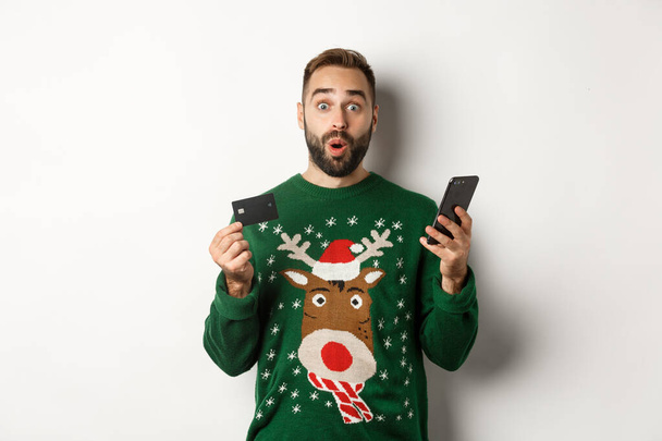 New Year, online shopping and christmas concept. Excited man using credit card and smartphone, standing over white background - Foto, Imagem