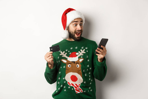 Online shopping and winter holidays concept. Man looking excited at mobile phone while buying in internet with credit card, wearing Santa hat, white background - Photo, Image