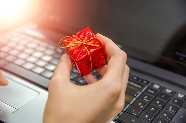 Kobieta trzyma w ręku małe czerwone pudełko z prezentami, z bliska - na tle laptopa, widok z tyłu. Zdjęcie z oświetleniem. Koncepcja Bożego Narodzenia, Nowy rok, święto - Zdjęcie, obraz