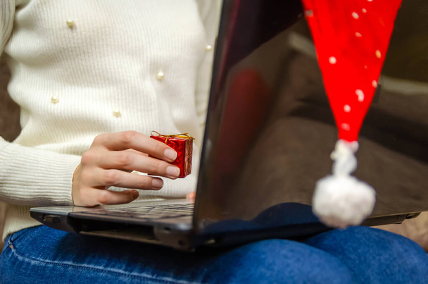 Woman in a white sweater holds a laptop on her lap with a Santa Claus hat hanging on it and holds a red gift box close-up in her hand. Concept of Christmas, New year, holiday - Valokuva, kuva