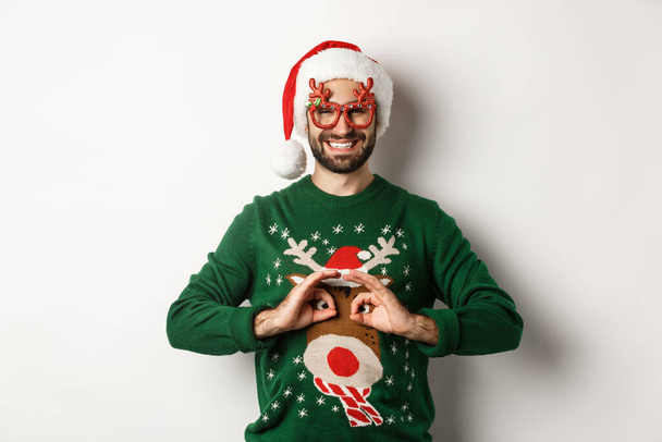 Різдвяні свята, концепція святкування. Щасливий хлопець в капелюсі і вечірні окуляри роблять смішний светр, стоячи на білому тлі
 - Фото, зображення