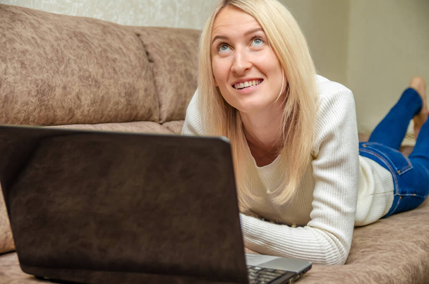 Jovem loira está deitada no sofá com um laptop, sorrindo e olhando para cima misteriosamente. Conceito de comunicação online, auto-isolamento - Foto, Imagem