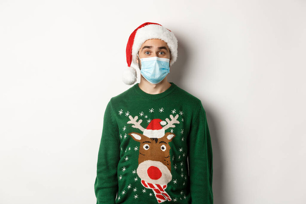 Christmas during pandemic, covid-19 concept. Young man in Santa hat and face mask celebrating New Year party with preventive measures, white background - Valokuva, kuva