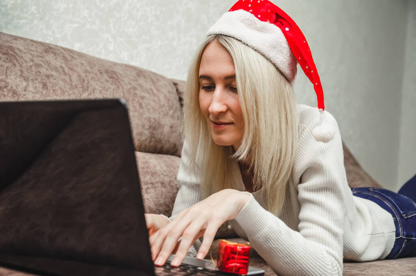 Jonge mooie blonde vrouw met een kerstmuts ligt op de bank met een laptop en typt een bericht. Kerstconcept, Nieuwjaar. Concept van online communicatie. - Foto, afbeelding
