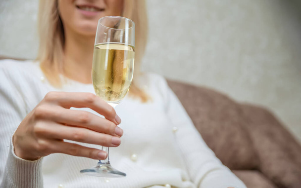 Young blonde woman in a white sweater with a glass of champagne is sitting on the sofa, copying the space. Focus on the glass - Photo, Image