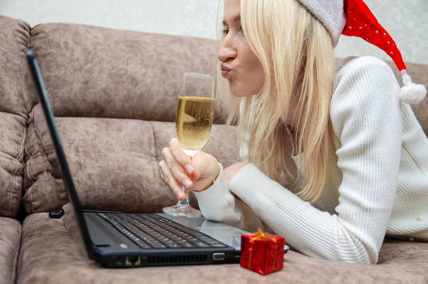 Jonge mooie blonde vrouw met een kerstmuts ligt op de bank met een laptop, een glas champagne met een vrolijke grimas op haar gezicht kijkend naar de laptop camera. Kerstconcept, Nieuwjaar. - Foto, afbeelding