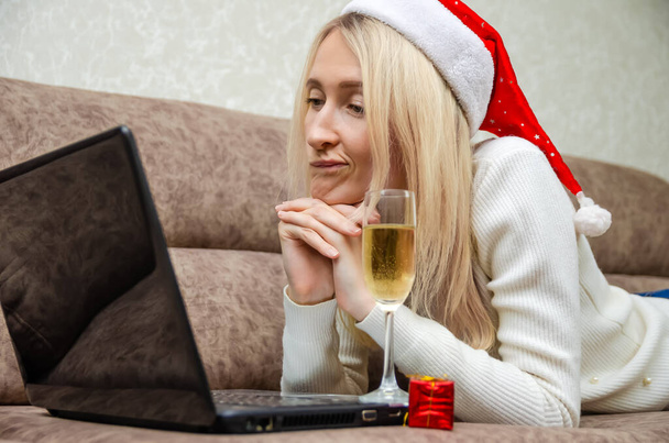 Młoda, piękna blondynka w kapeluszu Mikołaja leży na kanapie z laptopem, kieliszkiem szampana z grymasem na twarzy patrzącym w kamerę na laptopie. Koncepcja Bożego Narodzenia, Nowy Rok. - Zdjęcie, obraz