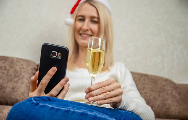 Young beautiful blonde woman in a Santa hat in a white sweater with a glass of champagne smiles and communicates via mobile phone. Concept of Christmas, New year, online communication - Foto, Imagen