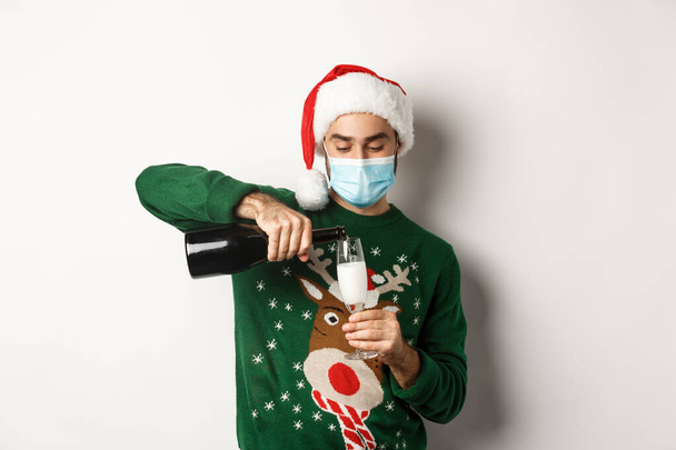 Concept of covid-19 and Christmas holidays. Happy guy in face mask and santa hat, celebrating New Year party and pouring glass of champagne, white background - Photo, image