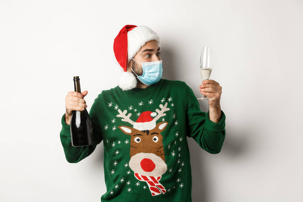 Concept of covid-19 and Christmas holidays. Man in medical mask and Santa hat enjoying glass of champagne, celebrating New Year, standing over white background - Photo, image