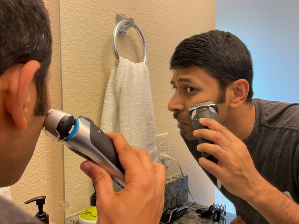 Закройте вид на человека, смотрящего в зеркало и использующего электробритву для бритья - Фото, изображение
