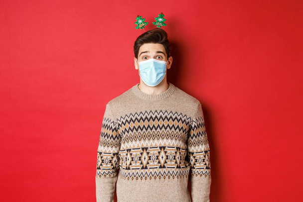 Noel konsepti, covid-19 ve sosyal uzaklık. Tıbbi maskeli neşeli genç adam ve yeni yıl kıyafetleri, salgın sırasında tatilleri kutlayan, kırmızı arka planda duran - Fotoğraf, Görsel