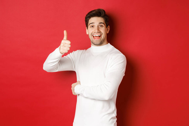 Concept of winter holidays, christmas and lifestyle. Portrait of cheerful, attractive man in white sweater, showing thumbs-up in approval, like good idea, standing over red background - Fotografie, Obrázek