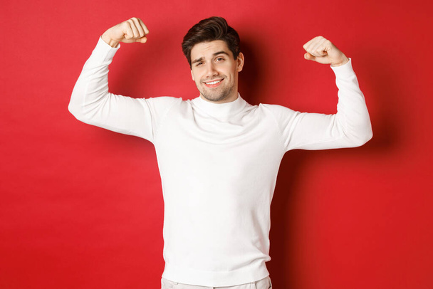 Portré mosolygós jóképű férfi fehér pulóver, hajlító bicepsz és kérkedett az erő, hencegő erős izmok edzés után, álló felett piros háttér - Fotó, kép