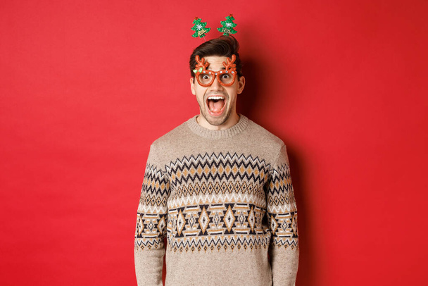 Şaşırmış ve heyecanlı yakışıklı bir adamın resmi, parti gözlüğü takıyor, Noel süveteri giyiyor, çenesini indiriyor ve kırmızı arka planda duran yeni yıl tanıtım teklifine bakıyor. - Fotoğraf, Görsel