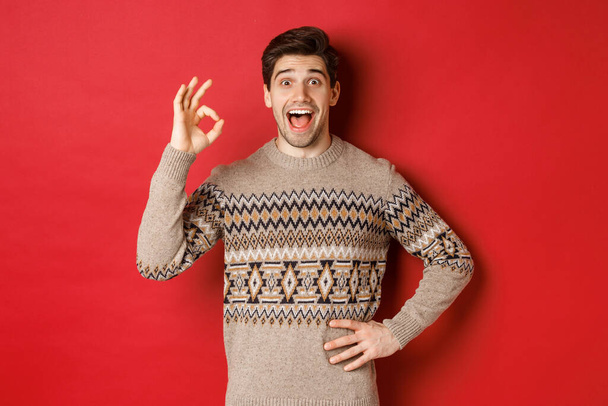 Концепция празднования Рождества, зимних праздников и образа жизни. Удивительный и счастливый человек в свитере xmas, показывающий хороший знак и смотрящий на что-то фантастическое на красном фоне - Фото, изображение