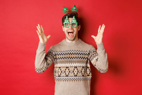 Imagem do cara caucasiano animado e surpreso em óculos de festa, camisola de Natal, levantando as mãos e fazendo grande anúncio, desfrutando de celebração do ano novo, fundo vermelho - Foto, Imagem