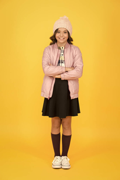 Fashionable girl looks like hipster. Small girl winter hat. Trendy schoolgirl wear jacket skirt and long socks. Clothes shop. Cool girl. Happy child yellow background. Warm clothes for cold season - Fotoğraf, Görsel