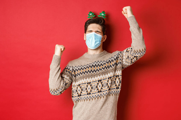 Concept of coronavirus, winter holidays and social distancing. Image of excited and happy man in medical mask and christmas clothing, raising hands up and celebrating new year, red background - Фото, изображение