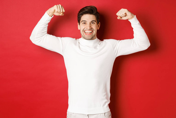 Retrato de hombre guapo sonriente en suéter blanco, bíceps flexionantes y jactancia con fuerza, músculos fuertes después del entrenamiento, de pie sobre fondo rojo - Foto, imagen