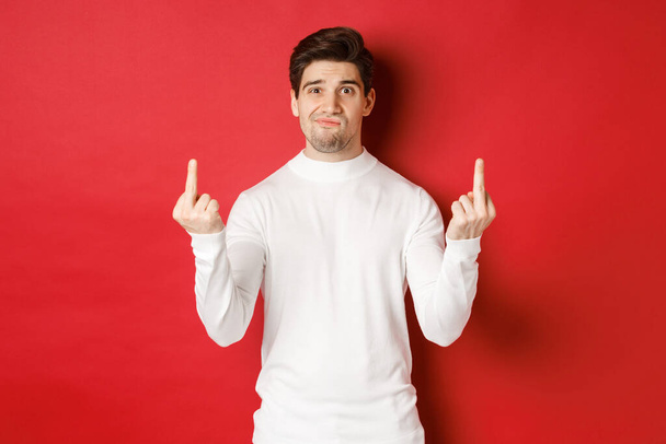 Image of pissed-off and distressed man telling to fuck off, showing middle-fingers and looking upset, standing over red background in white sweater - Fotografie, Obrázek