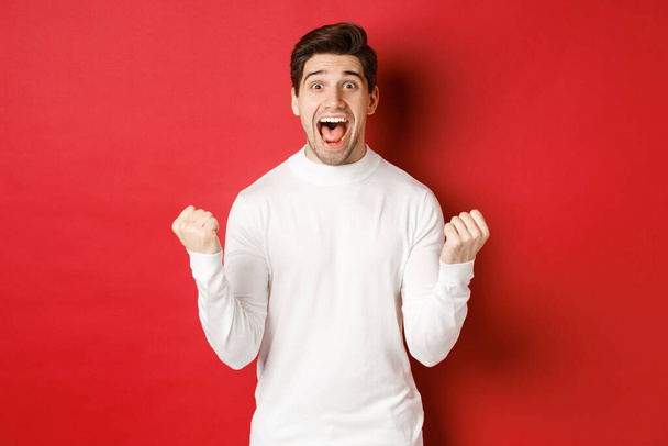 Image of happy good-looking man in white sweater, winning something, making fist pump and smiling amazed, celebrating victory, standing over red background - Photo, Image