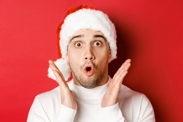 Close-up of amazed handsome man in santa hat and white sweater, raising hands and looking impressed, standing over red background - Φωτογραφία, εικόνα