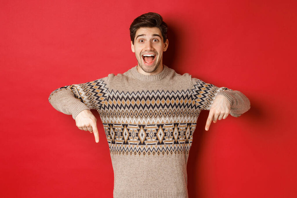 クリスマスのお祝い、冬の休日やライフスタイルの概念。興奮した白人男性モデルでxmasセーター,指差し指と発表を行います,広告を表示,赤の背景 - 写真・画像