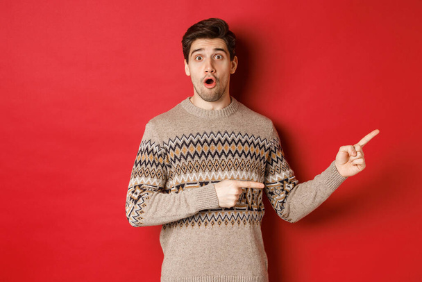クリスマスのお祝い、冬の休日やライフスタイルの概念。素晴らしいですハンサムな男でxmasセーター指して指右にロゴ,示す広告,立って上赤背景 - 写真・画像