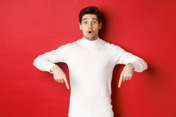 Image of surprised attractive guy in white sweater, showing announcement, pointing fingers down and looking amazed, standing over red background - 写真・画像