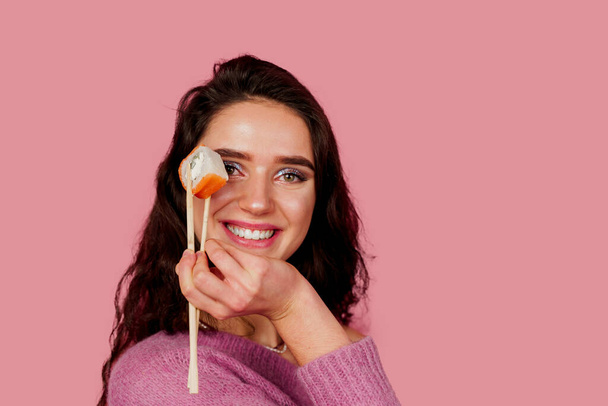 Happy girl with sushi isolated on pink background. Young girl is holding sushi by chopsticks, smiling, having fun and wanting to eat one piece of roll philadelphia. Advert for sushi delivery. - Photo, image