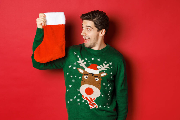 Εικόνα του έκπληκτος και διασκεδαστικός άνθρωπος σε πράσινο πουλόβερ, κοιτάζοντας χριστουγεννιάτικη κάλτσα με δώρα και χαμογελώντας, στέκεται πάνω από το κόκκινο φόντο - Φωτογραφία, εικόνα