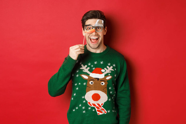Πορτρέτο του διασκεδάζει όμορφος άντρας σε χριστουγεννιάτικο πουλόβερ, κρατώντας αστεία μάσκα κόμμα και χαμογελώντας, γιορτάζει τις χειμερινές διακοπές, στέκεται πάνω από το κόκκινο φόντο - Φωτογραφία, εικόνα